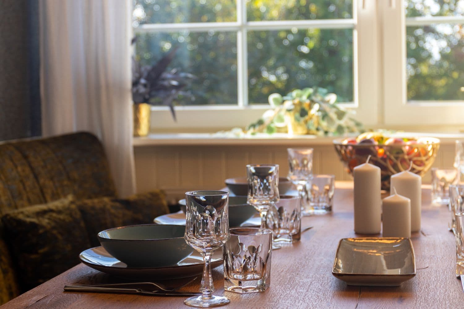 Laid dining table in backlight with crystal glasses, candles and a fruit bowl in front of the window