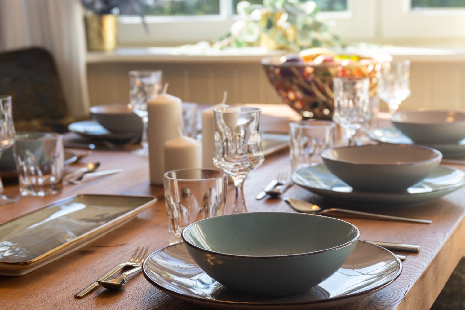 Grey-blue ceramic dishes, glasses and cutlery on the wooden table, with candles and a fruit bowl behind