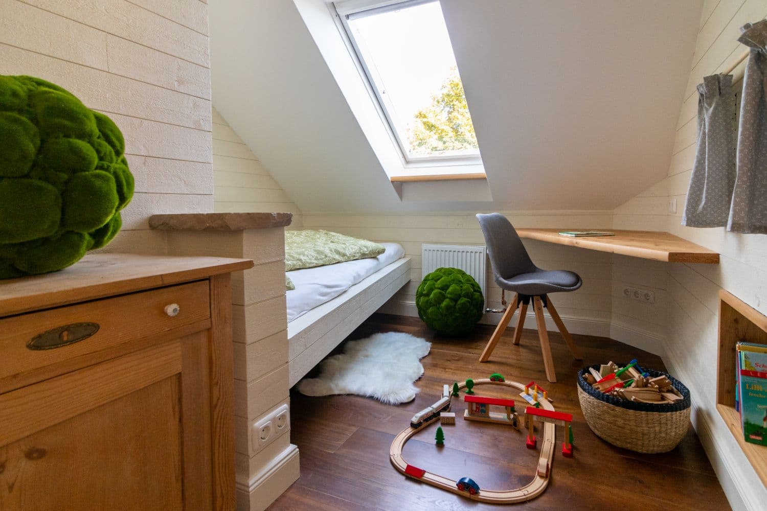 Children's room under the sloping ceiling with a bed, desk, wooden train set on the floor and a roof window