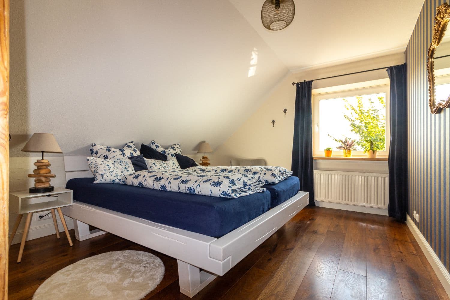 Bedroom with a white double bed, blue bed linen with a flower pattern, sloping ceiling and striped wallpaper