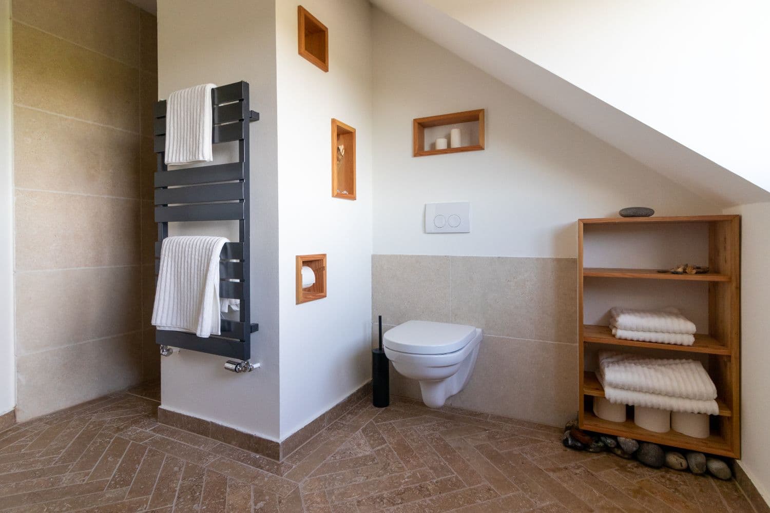 Bathroom with wall-hung toilet, black heated towel rail, wooden shelf with towels and herringbone floor