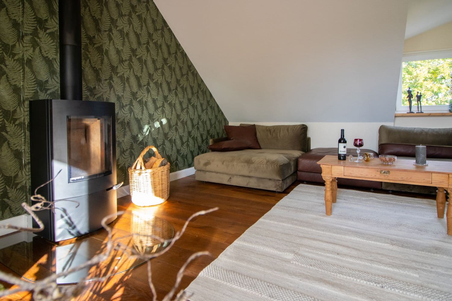 Living room with a black wood-burning stove in front of fern-green wallpaper, a basket of firewood, sofa set and coffee table under the sloping ceiling