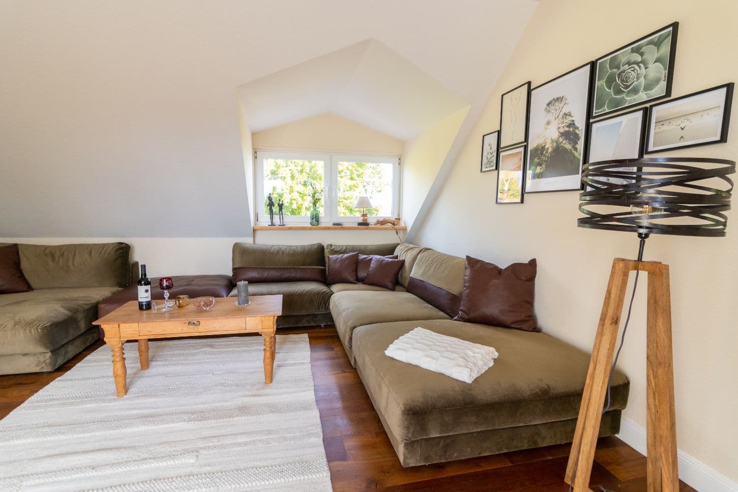 Olive green corner sofa with brown leather cushions in front of the roof window, a wooden table on a light rug and framed pictures on the wall