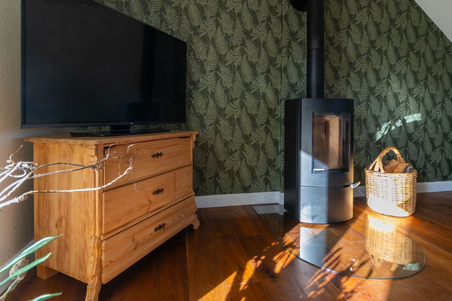 Television on an old wooden chest of drawers next to the wood-burning stove on its glass plate, with the log basket beside it