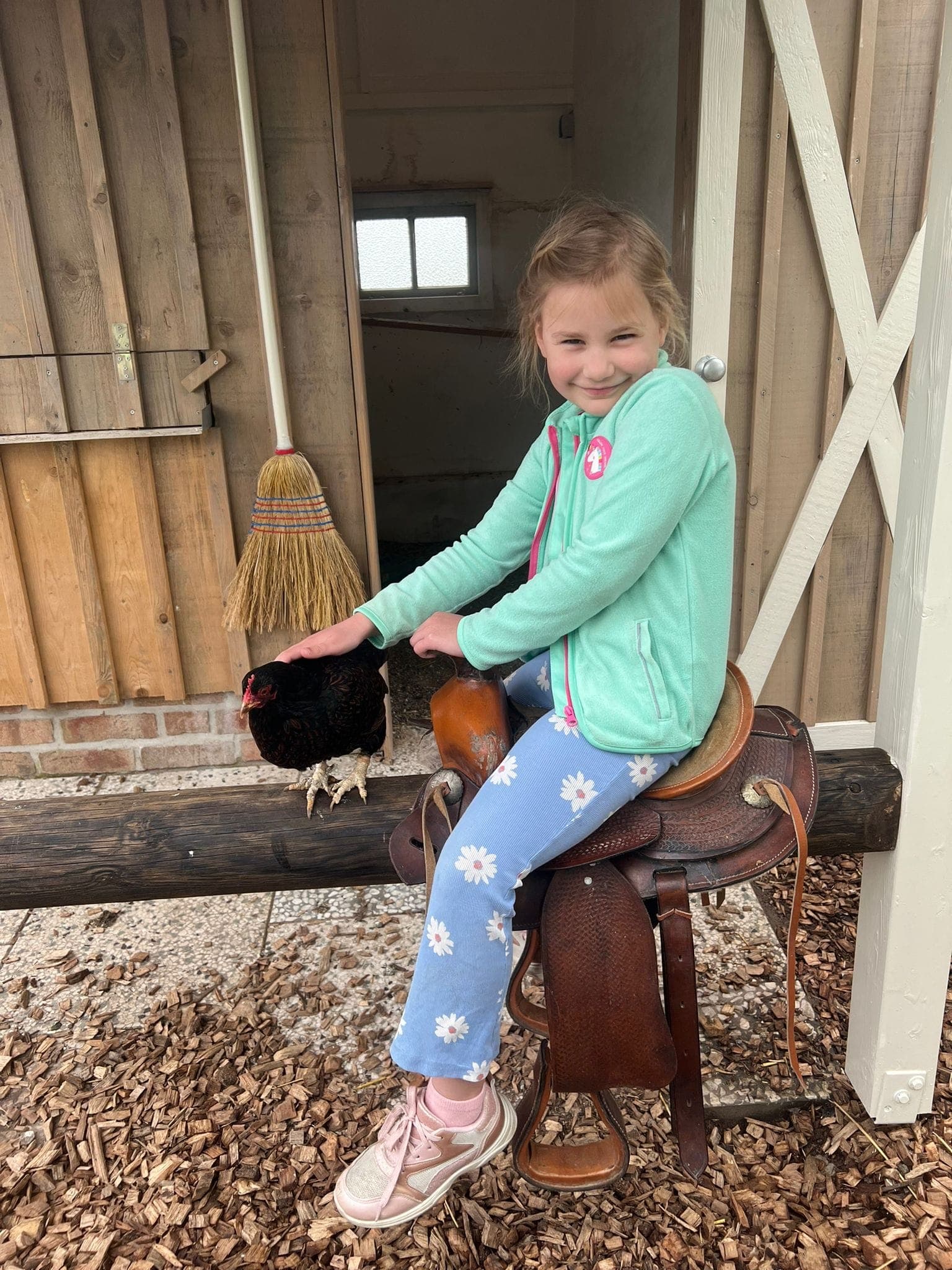 Kind in grüner Fleecejacke auf einem Westernsattel, daneben eine schwarze Henne vor dem Hühnerstall